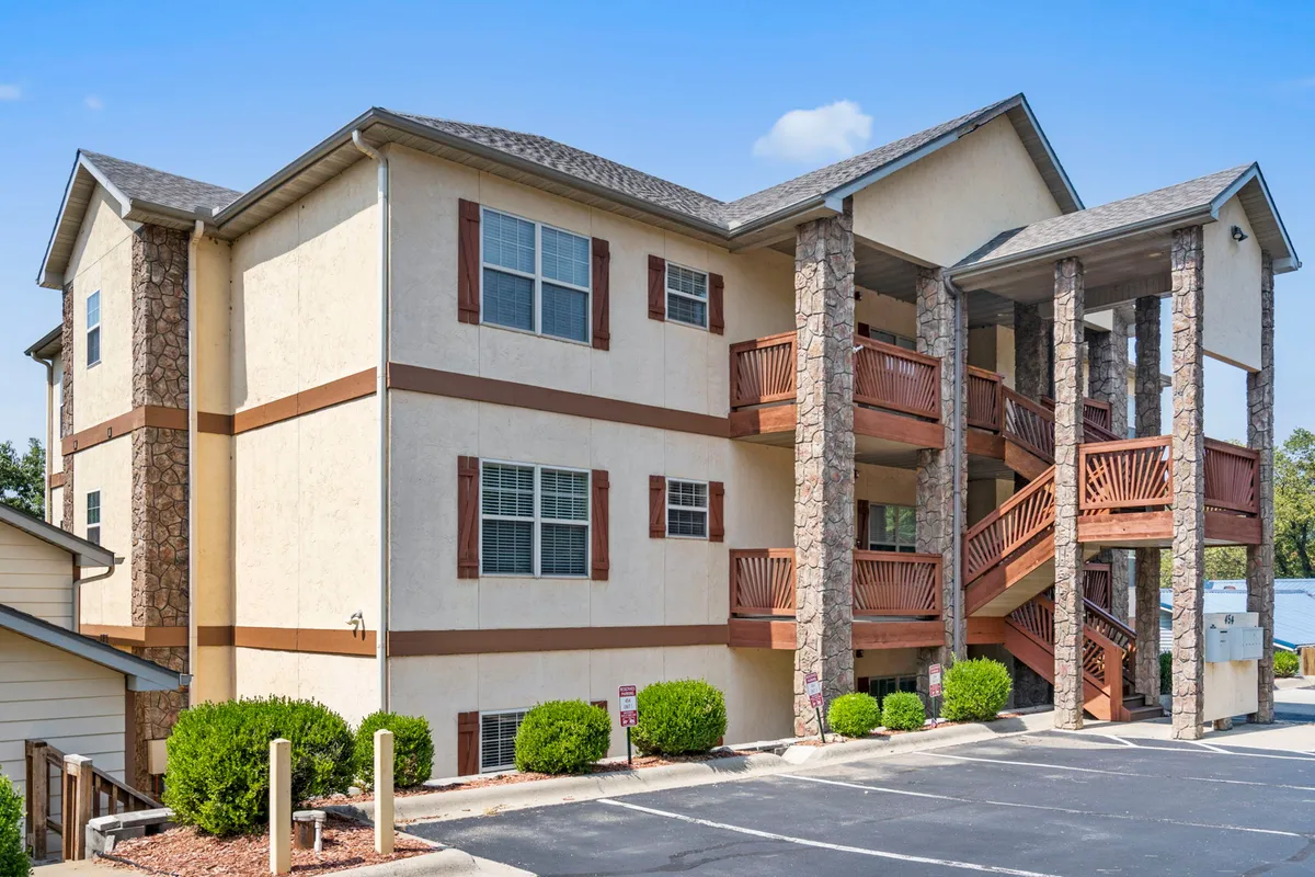 The exterior of the property is showcased, featuring stone columns, wooden balconies, and windows with shutters. Lush landscaped bushes and a spacious yard add to the inviting atmosphere, while balconies suggest opportunities for outdoor relaxation or socializing.