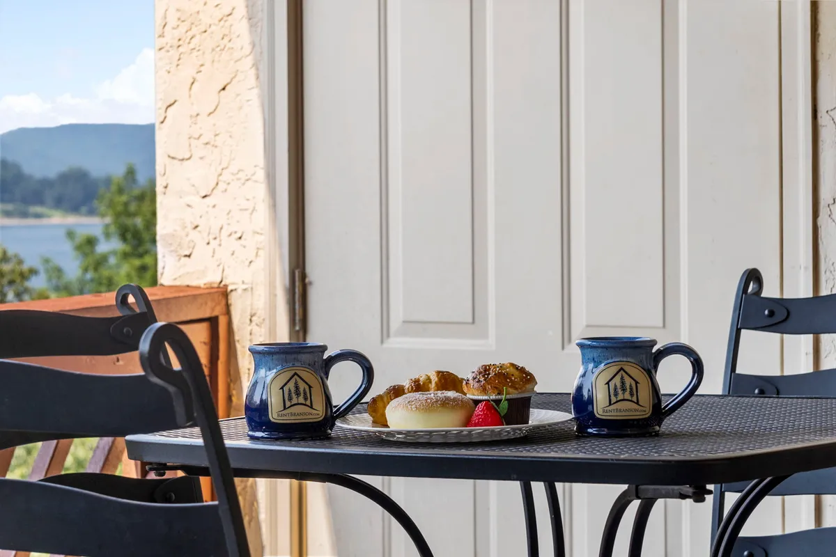 A relaxed outdoor breakfast scene with two coffee mugs, a plate of pastries, and a wide landscape view. The setting captures the essence of enjoying a meal or coffee while soaking in the natural beauty surrounding the home.