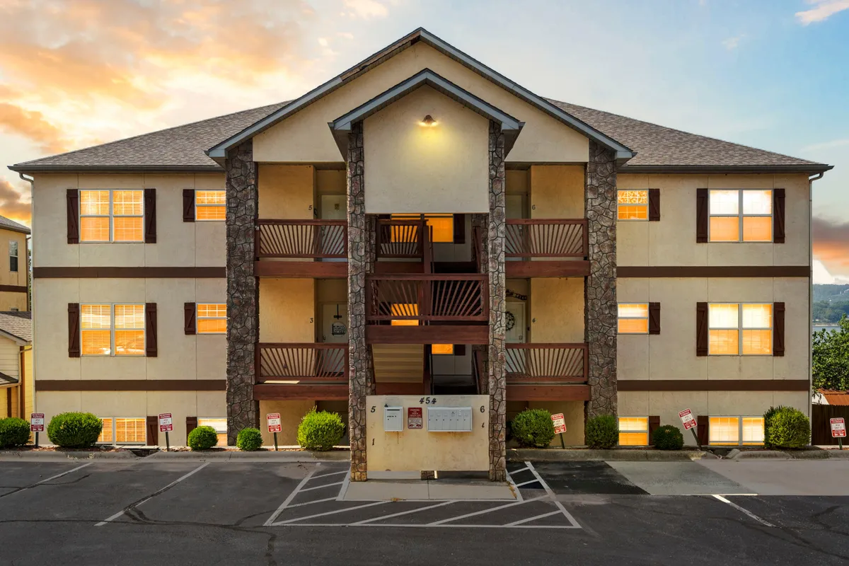 A welcoming exterior entrance with stone accents, glowing windows, and balconies. The soft evening light hints at community life, social balcony gatherings, and relaxing after a day of lakeside activities.