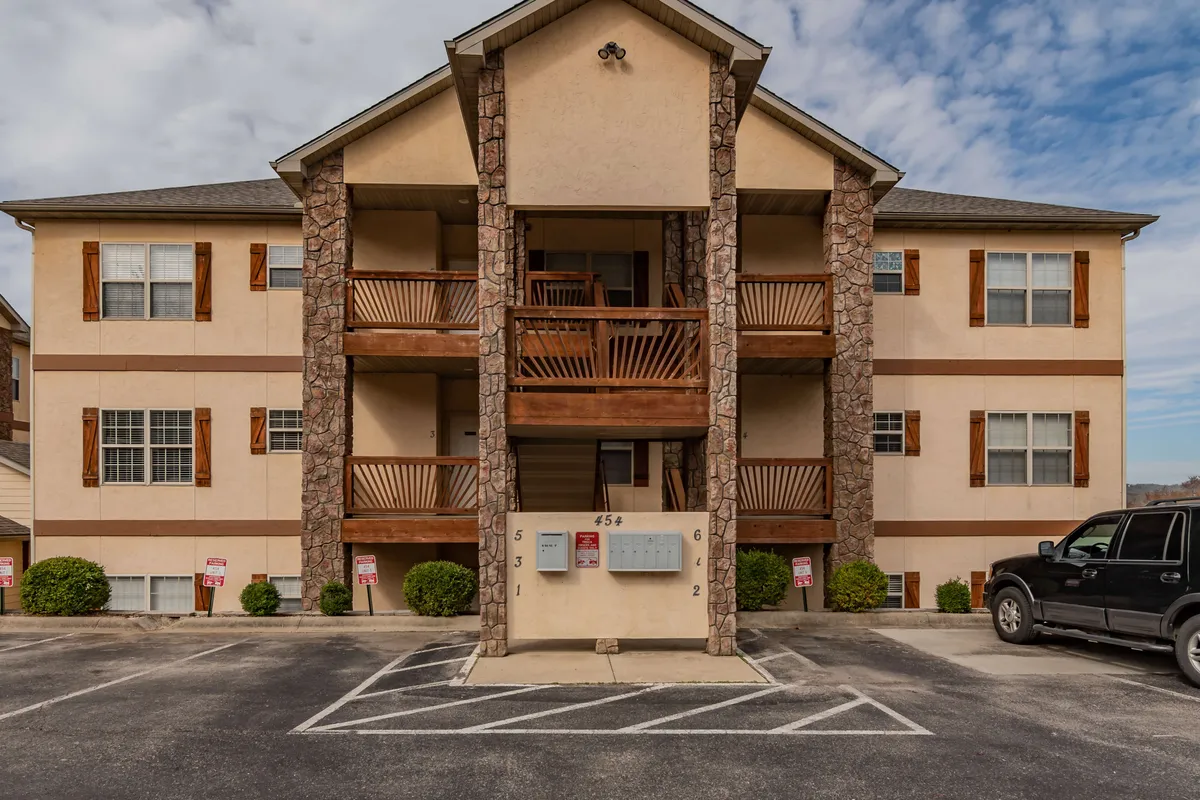 The image showcases the main entrance of the property, featuring a stone facade, wooden balconies, and a parking area with a vehicle. The inviting scene highlights communal outdoor spaces and the property's welcoming presence.