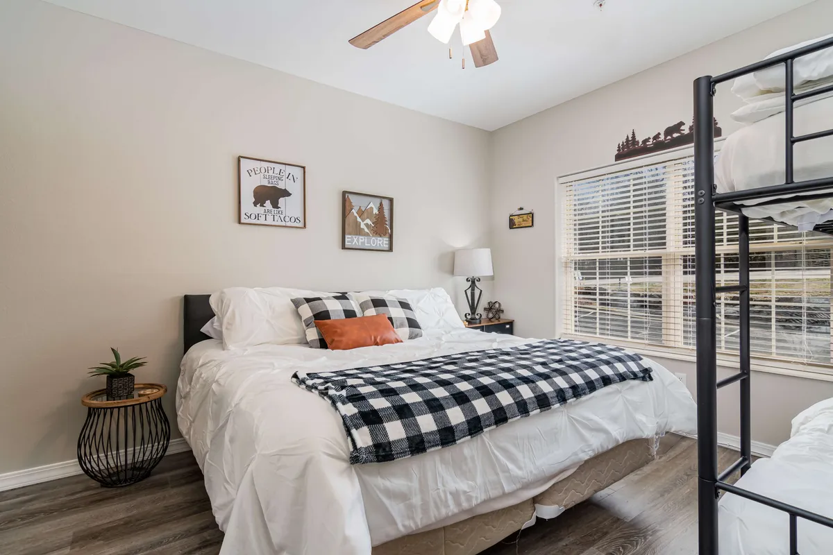 A family-friendly bedroom with a king bed adorned with a plaid blanket and an adjacent twin bunk bed, complemented by cheerful wall art and a nightstand with a lamp. The room feels spacious and adaptable for families or groups.