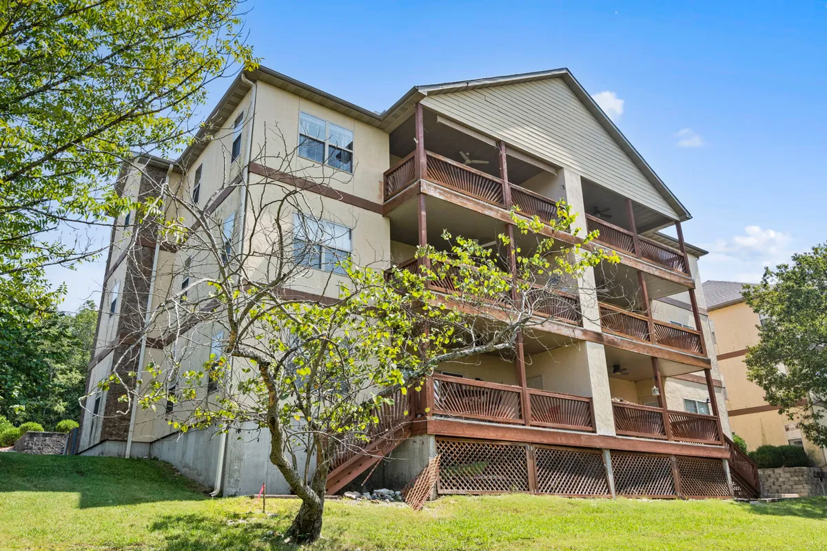 A broad exterior view of a three-story building with wooden balconies and a well-kept green lawn, set against a backdrop of natural greenery. The scene conveys a sense of community, privacy, and the option to enjoy outdoor relaxation or activities just outside the home.