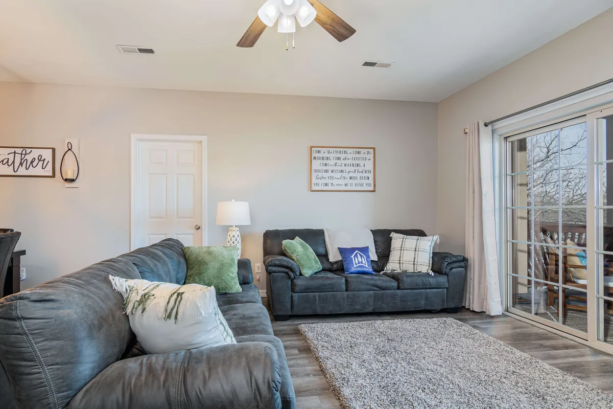 A cozy living room with plush sofas, decorative pillows, a standing lamp, artwork on the wall, and a sliding glass door that opens to a balcony. The atmosphere is warm and welcoming, perfect for relaxing, socializing, or enjoying family movie nights.
